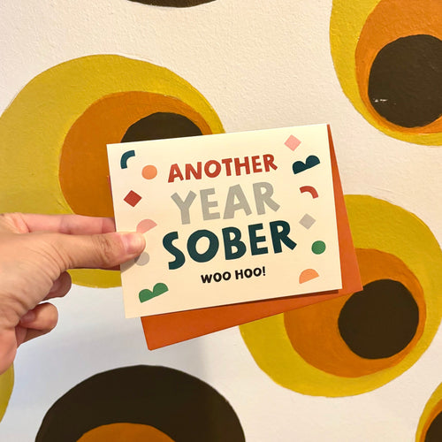 Hand holding a card with 'Another Year Sober' text against a patterned background