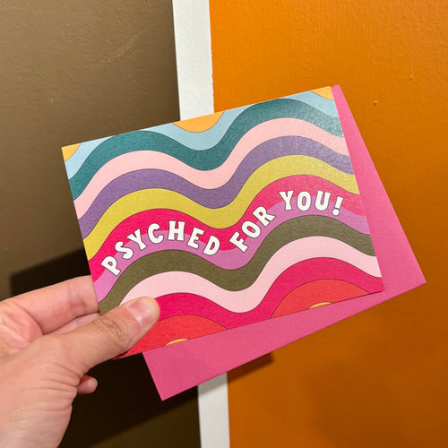 Colorful card with 'PSYCHED FOR YOU!' text held by a hand against a brown and orange wall.