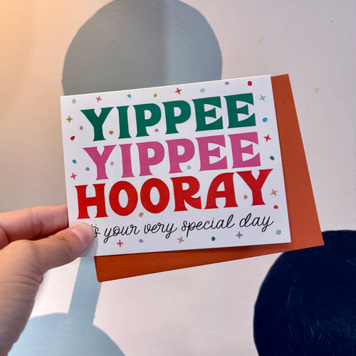 Hand holding a card with 'Yippee Yippee Hooray' text against a neutral background