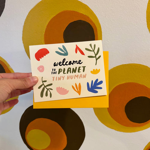 Hand holding a card with 'welcome to the planet tiny human' text against a patterned background