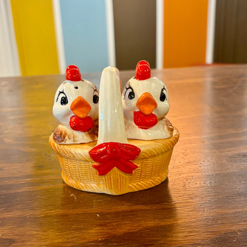 Decorative salt and pepper shakers shaped like chickens in a basket on a wooden table.
